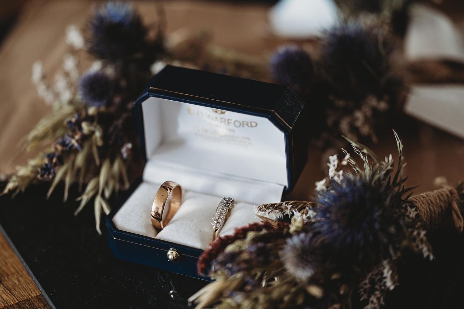 Wedding rings nestled in a white box surrounded by dried purple flowers and botanical elements.