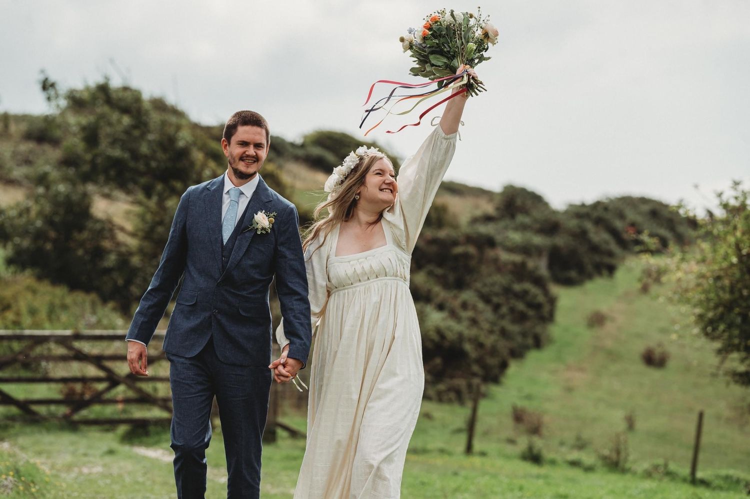Newlyweds walking through green countryside with bride holding bouquet triumphantly.