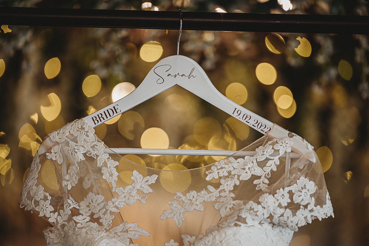 A delicate lace wedding dress hangs on a personalized hanger against a golden bokeh background.