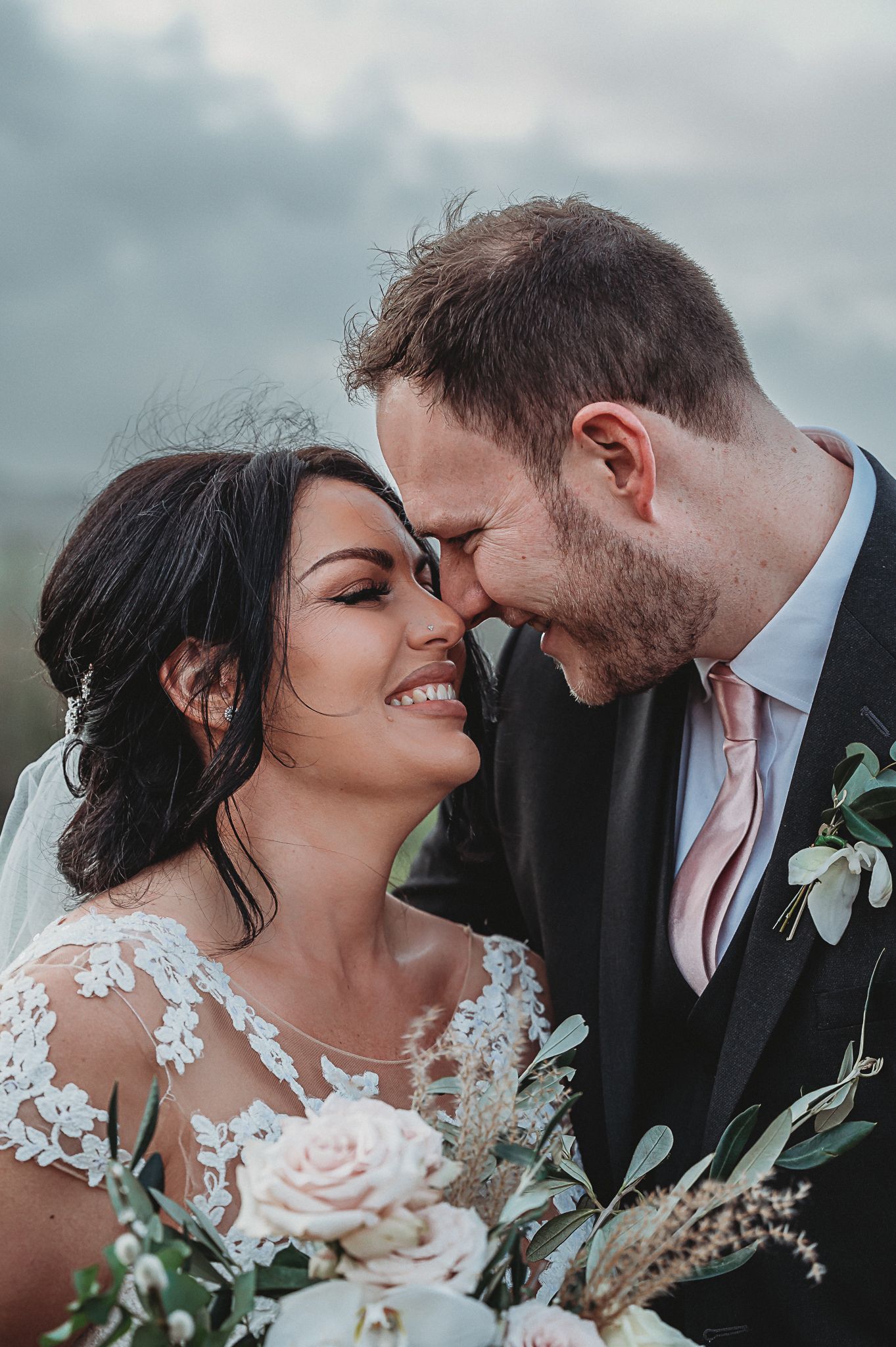 A romantic wedding portrait shows a couple sharing an intimate moment with soft pink roses and greenery bouquet.
