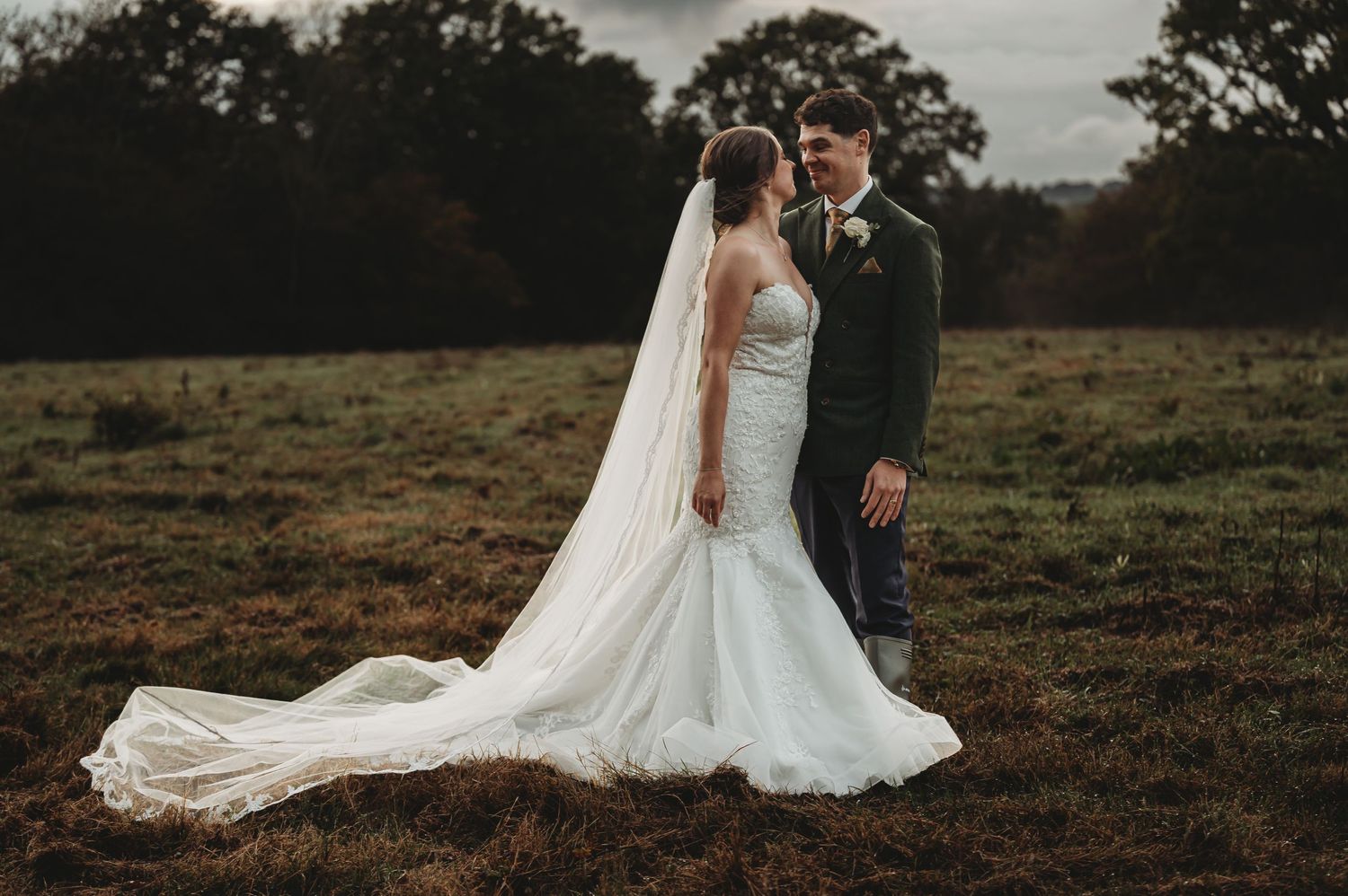 Bride in flowing white dress and groom embrace in an autumn field with dramatic lighting.