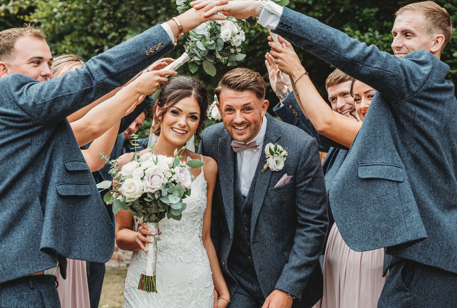 Wedding party forms arch with arms over couple in matching grey suits and white bouquet.
