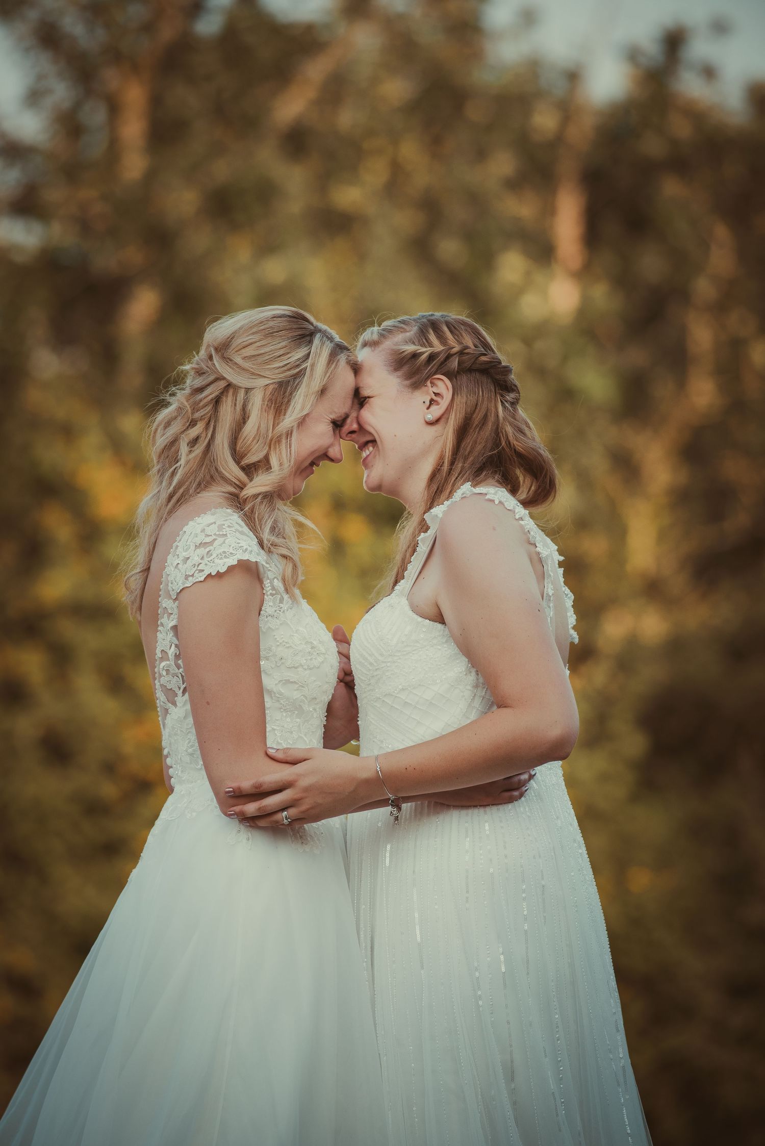 Two brides in matching white gowns share an intimate moment against a golden sunset background.