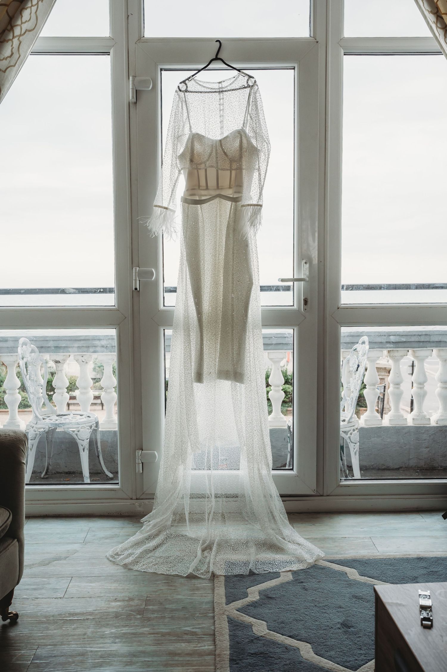 Elegant white wedding dress with sheer details hanging in front of french doors with balcony view.