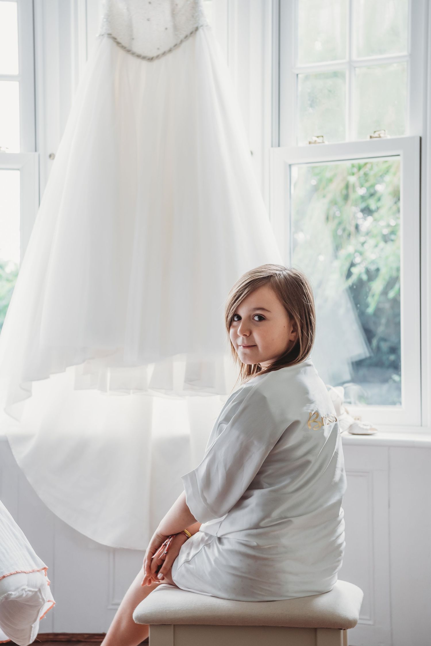 Wedding dress hangs by window while figure in white robe sits on bench below.