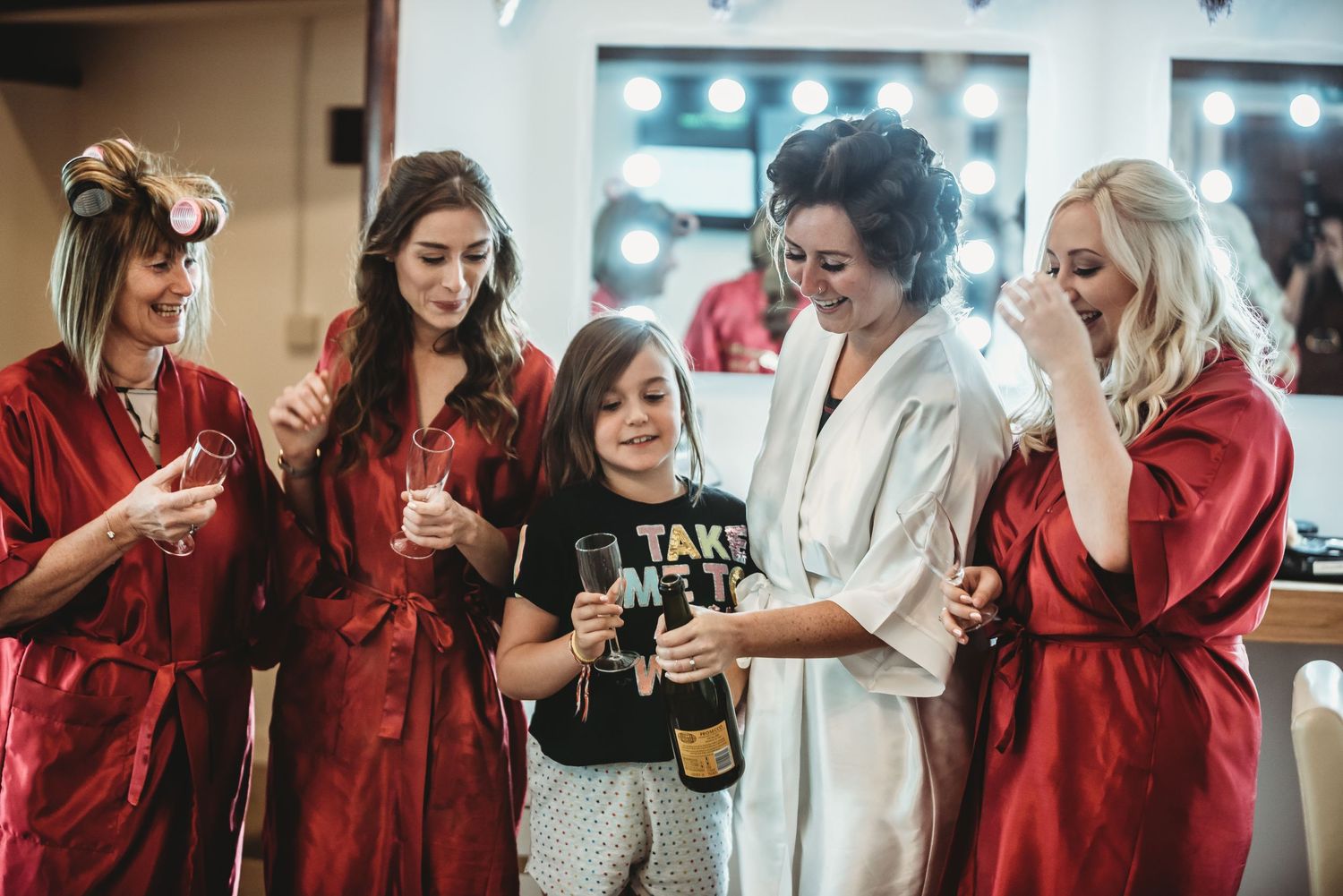 Group wearing red robes celebrate with champagne during wedding preparations.