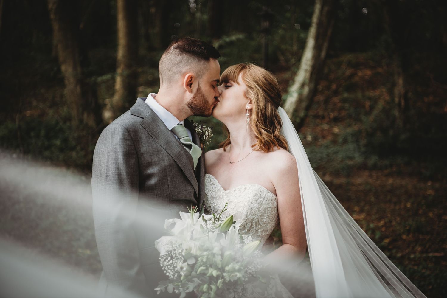 Intimate forest wedding portrait through soft focus with white bouquet and bridal veil.