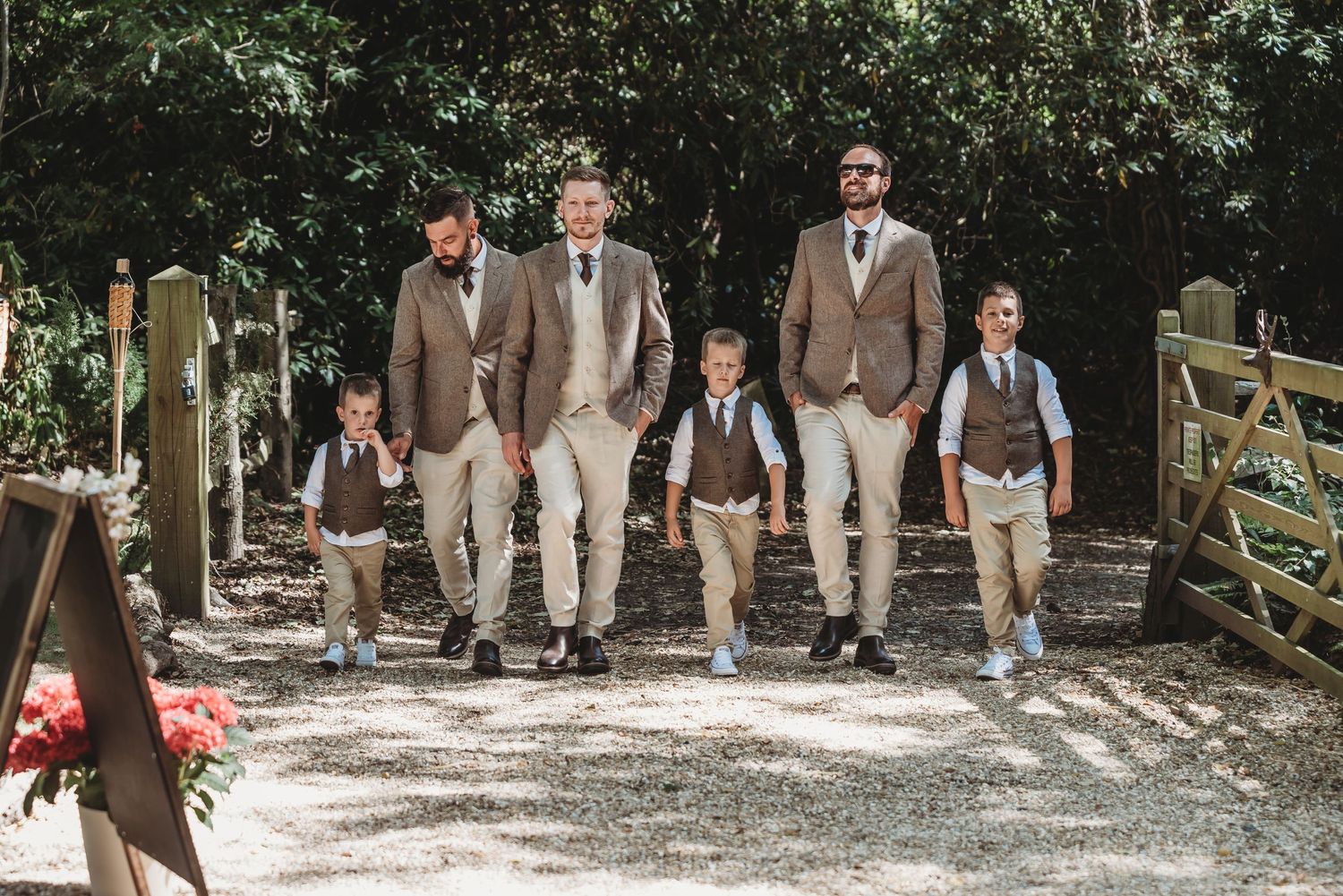 Groomsmen in light brown suits walk along a wooded path near a rustic gate.