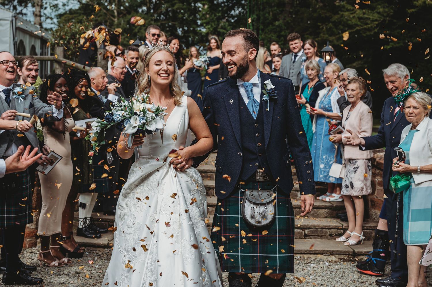 Confetti fills the air around a couple in traditional Scottish wedding attire surrounded by guests.