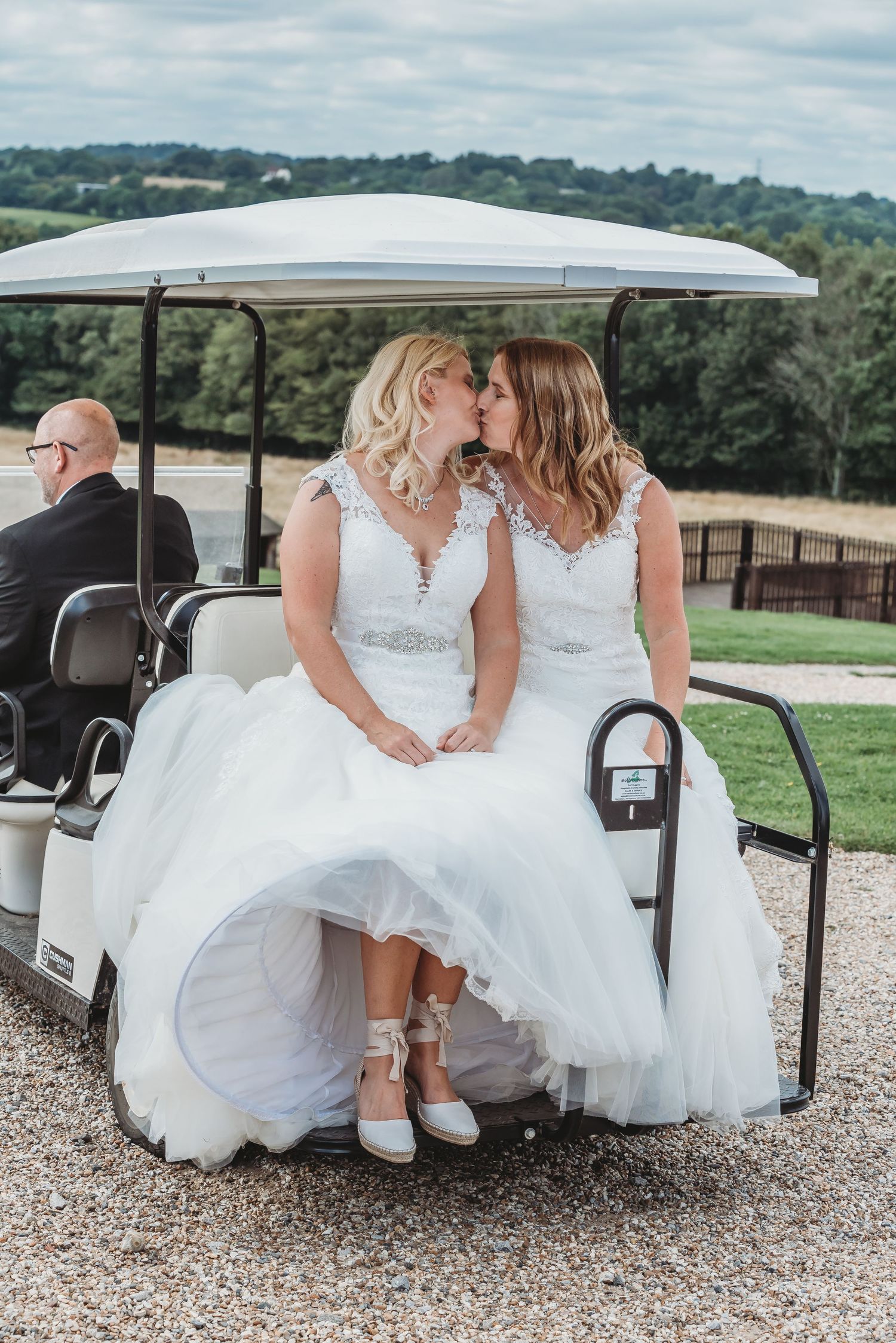 Two brides in white dresses share a sweet moment while sitting in a golf cart overlooking scenic hills.