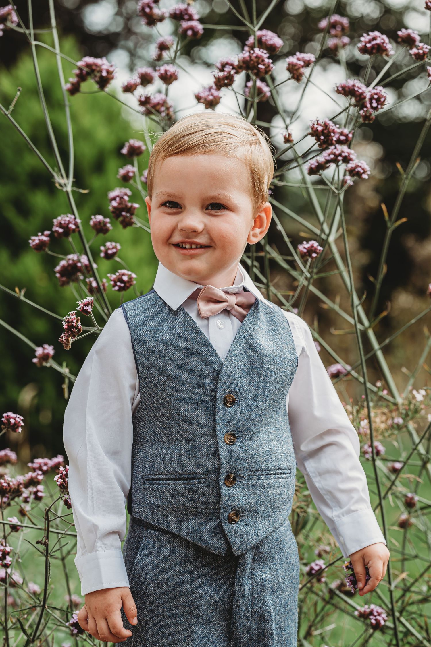 Young child in grey vest and bowtie smiles while standing among purple flowering plants.