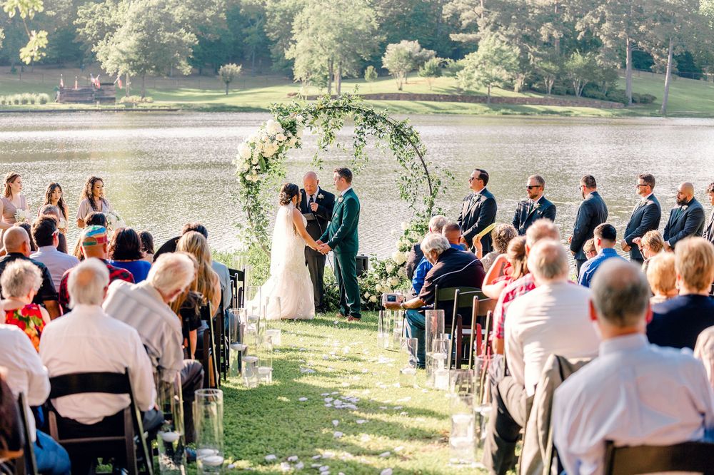 Outdoor wedding ceremony under a floral arch with guests seated on white chairs overlooking a scenic landscape.