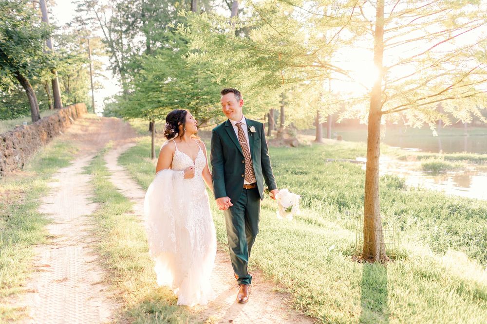 A couple walks hand in hand along a sunlit dirt path with a scenic lake view in the background.