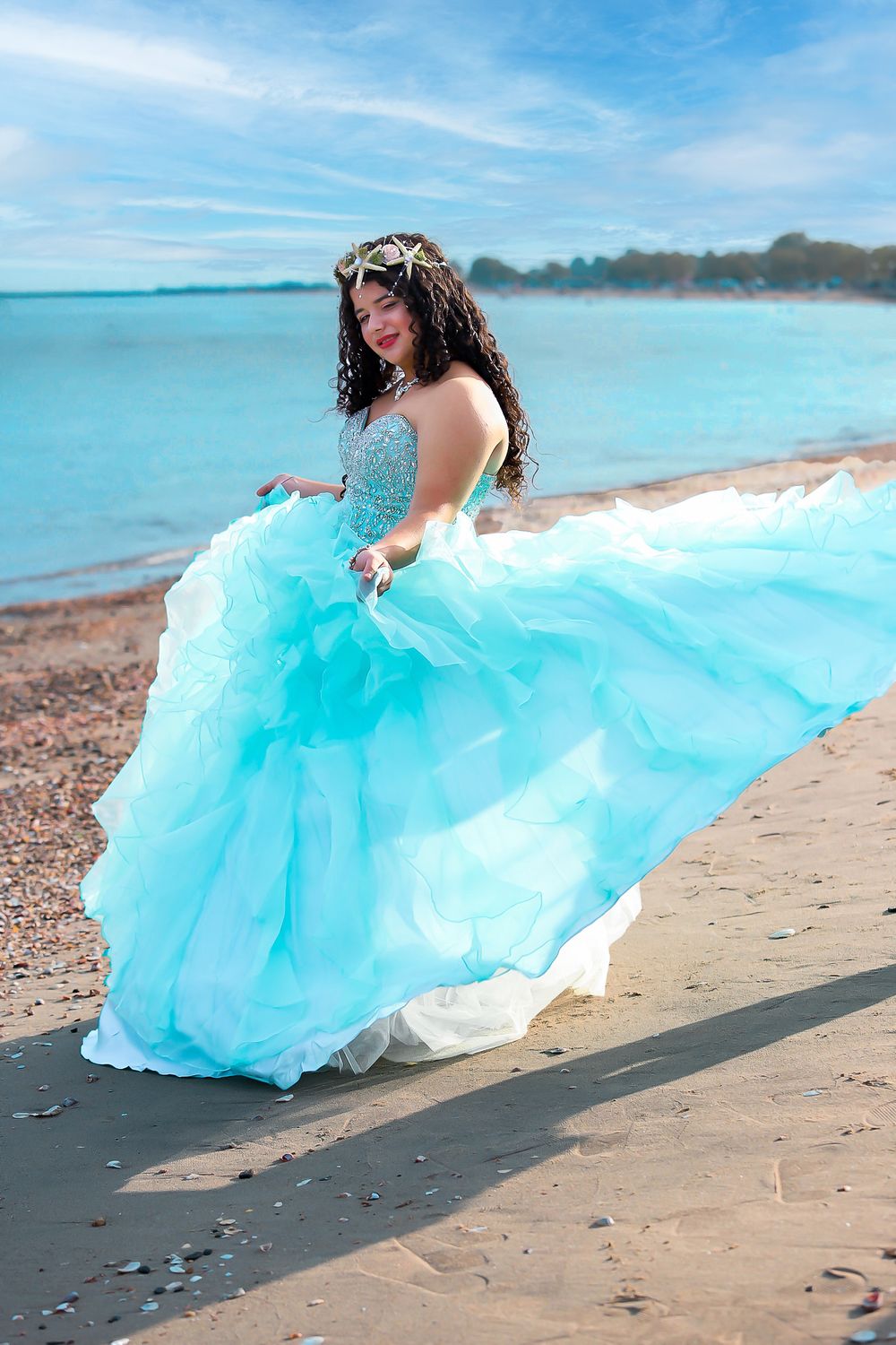 A flowing turquoise quinceanera gown billows in the breeze on a scenic beach with blue waters in the background.