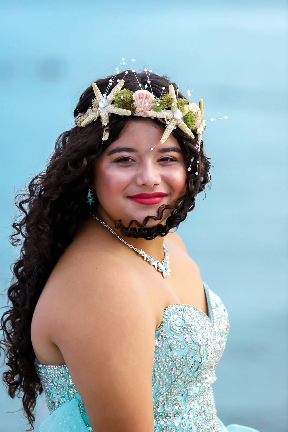 A beach-themed portrait with seashell crown and aqua blue formal dress against ocean backdrop.