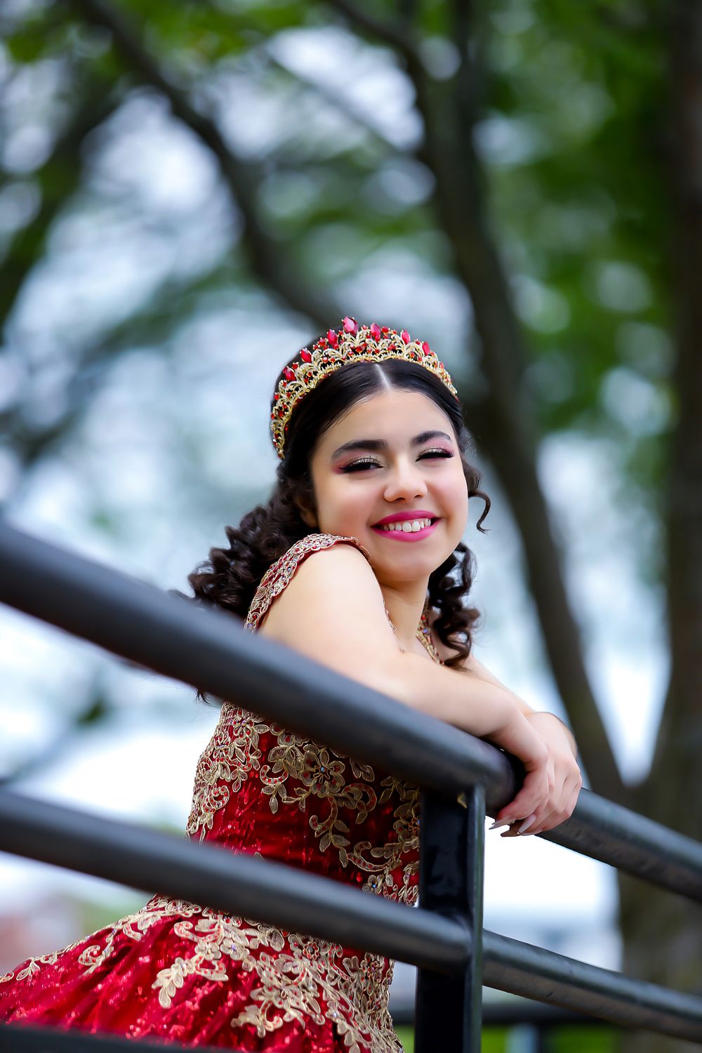 A celebration portrait in a red and gold embellished gown with a crown against a natural outdoor backdrop.