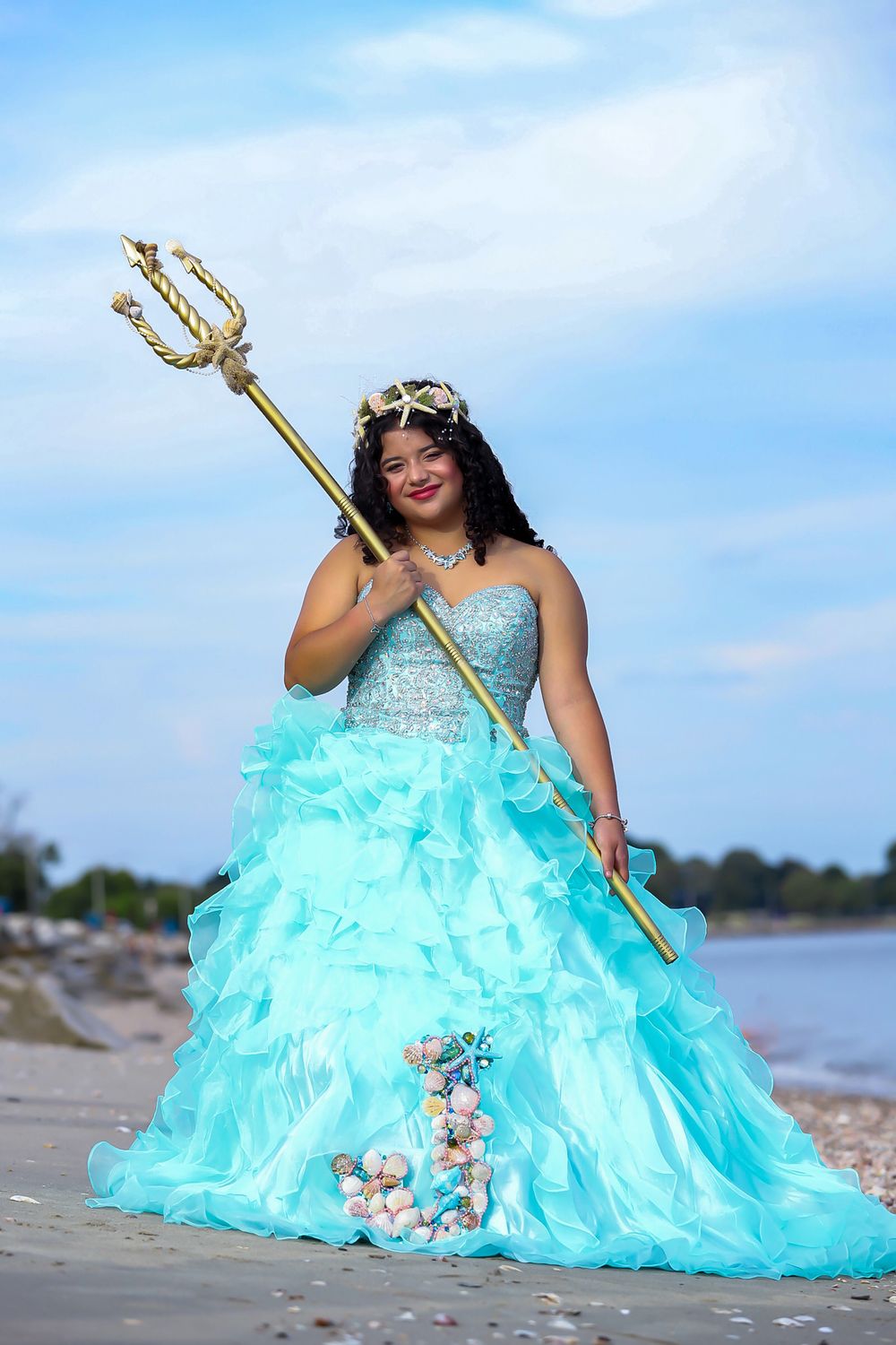 A whimsical beach portrait in aqua ball gown holding a golden trident against coastal scenery.