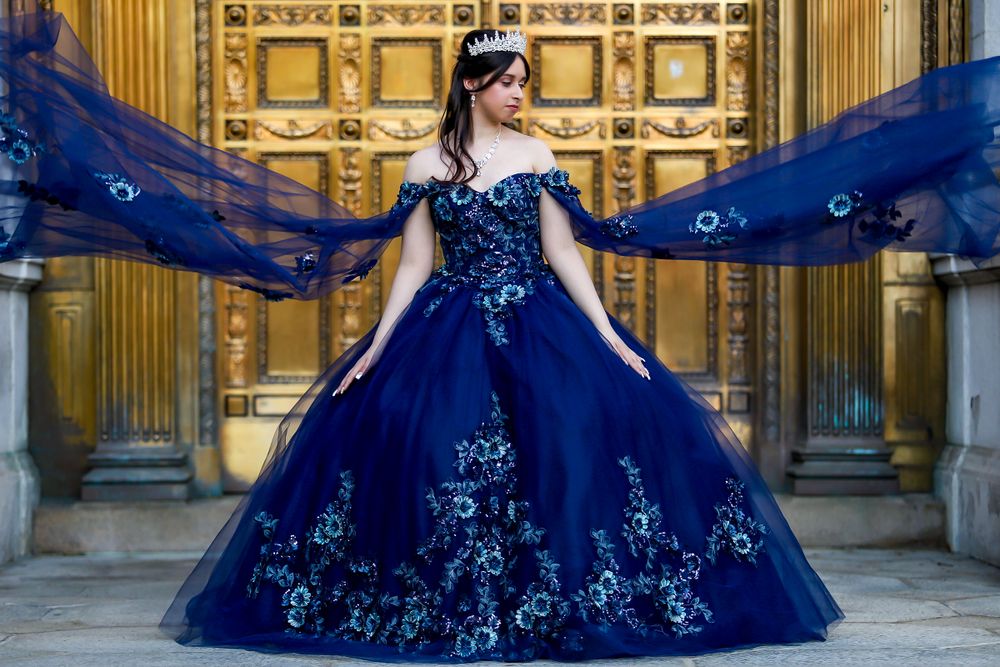 A dramatic navy blue ball gown with flowing cape creates an elegant silhouette against golden architecture.