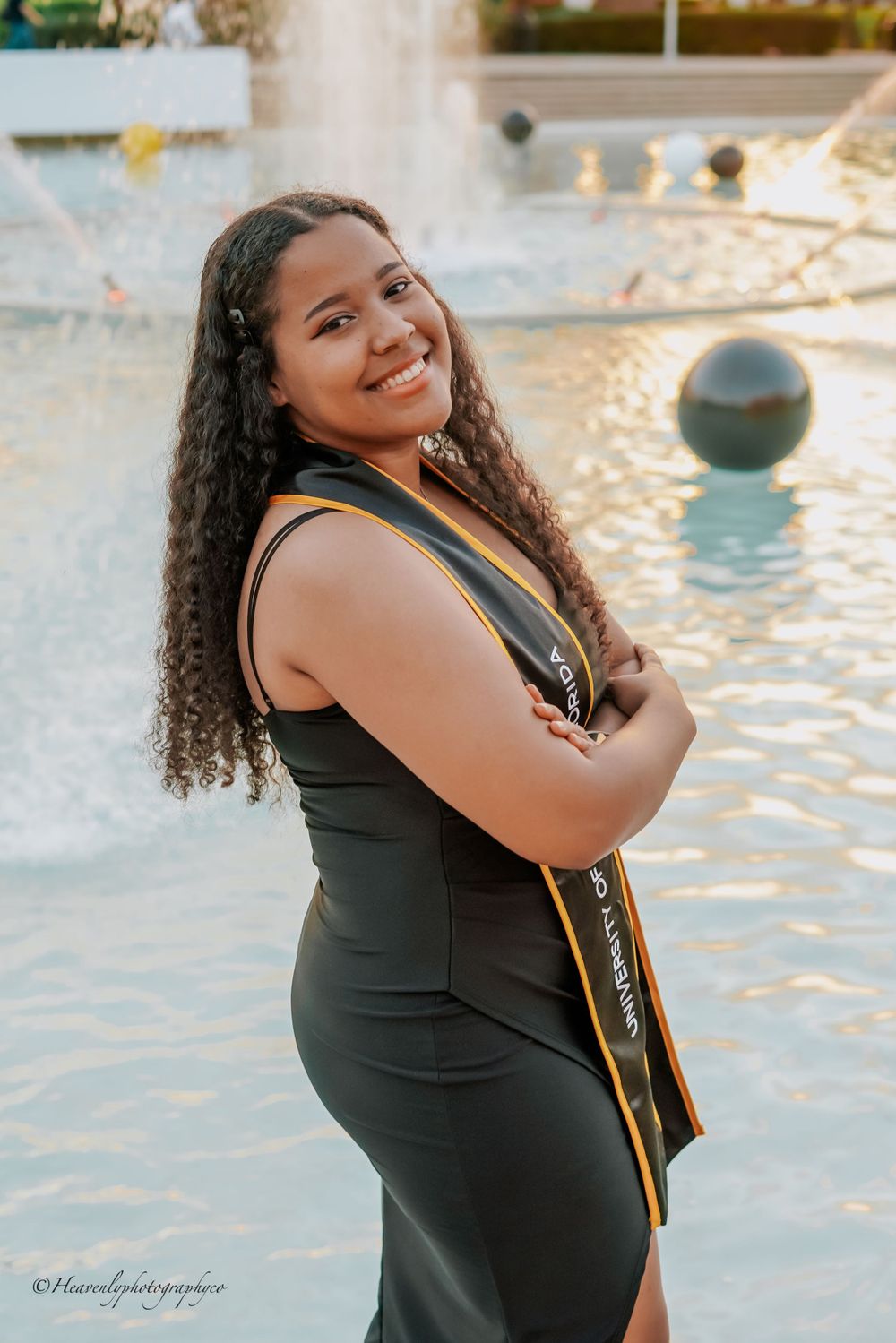 Graduate in black dress and gold stole poses joyfully by water fountain.
