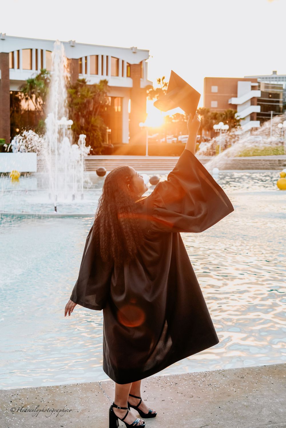 Silhouette of graduate in flowing gown poses dramatically by fountain at sunset.