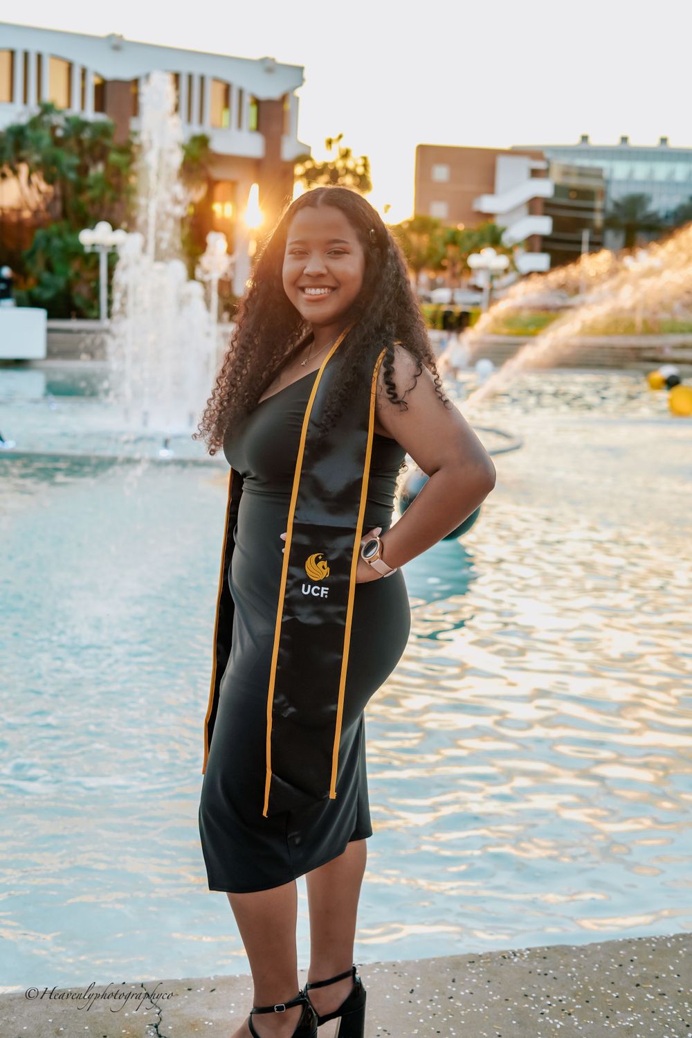 Graduate in black gown and gold stole poses by fountain during sunset.