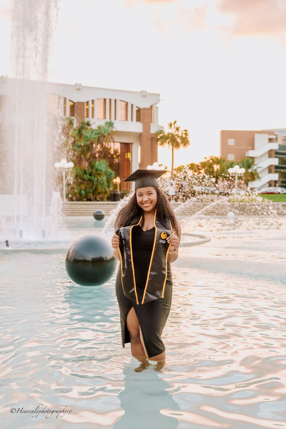 Graduate wading through campus fountain water in graduation attire during sunset.