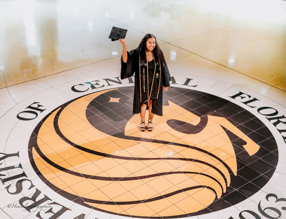 Graduate standing on university seal in graduation regalia holding cap up.
