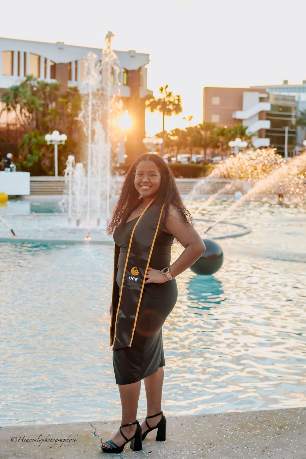 Graduate in black gown poses confidently by fountain during golden hour photoshoot.