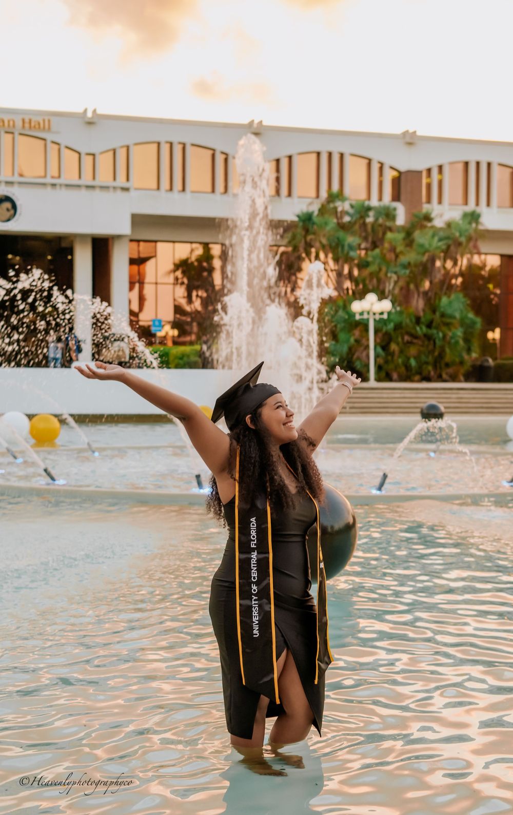 Graduate celebrating in a fountain wearing cap and gown with arms raised during golden hour.