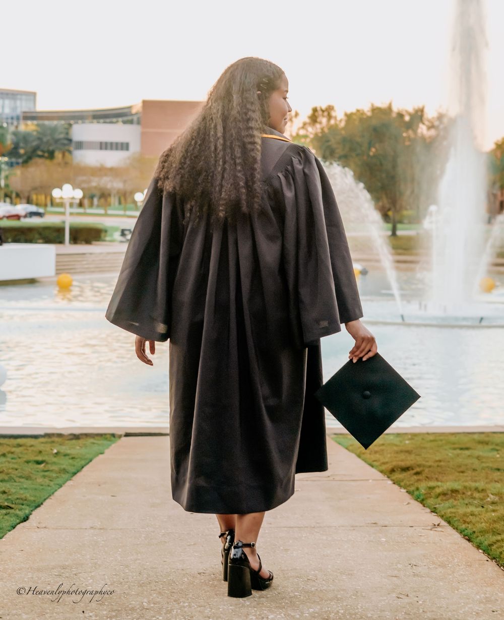 Graduate in black cap and gown walking away while holding diploma near fountain at sunset.