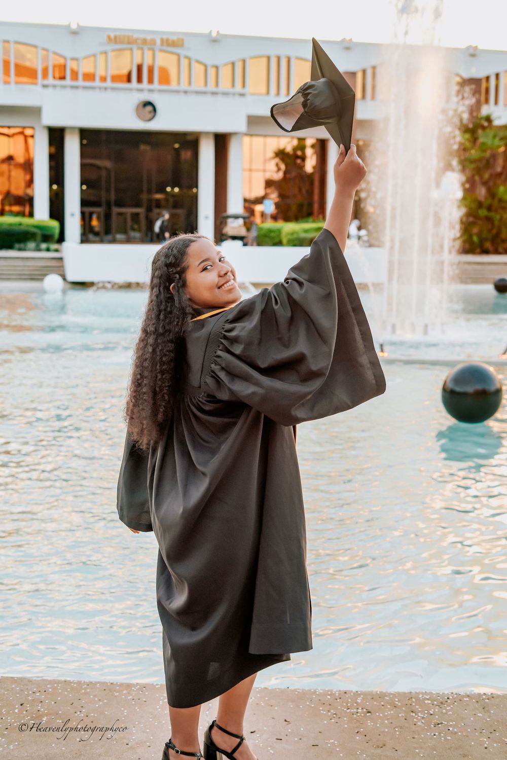Graduate celebrates with arms raised in black gown by fountain during sunset.