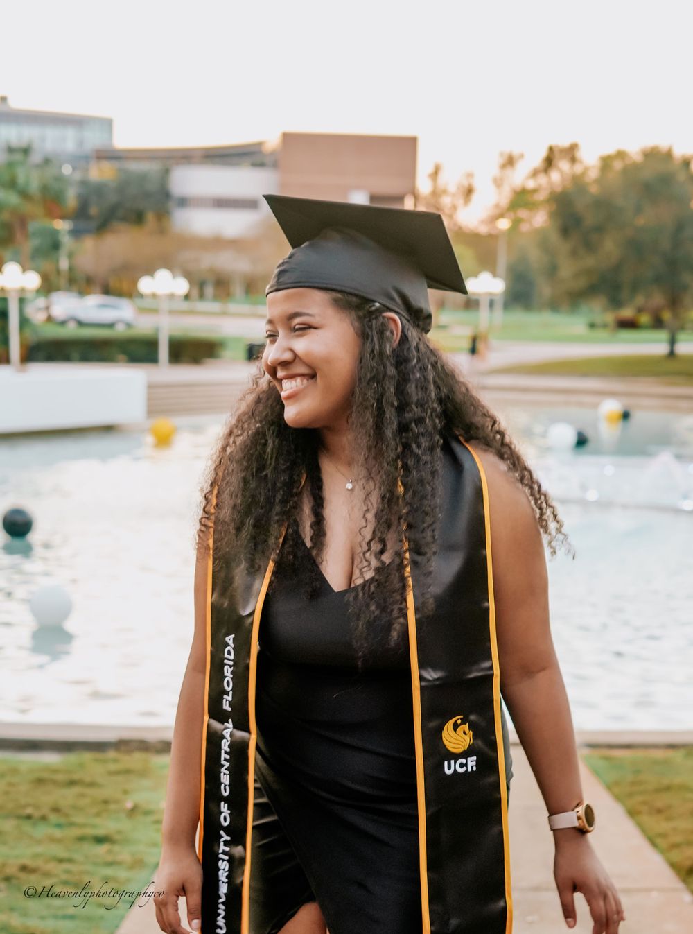 Graduate in black graduation gown and cap smiling outdoors near a fountain.