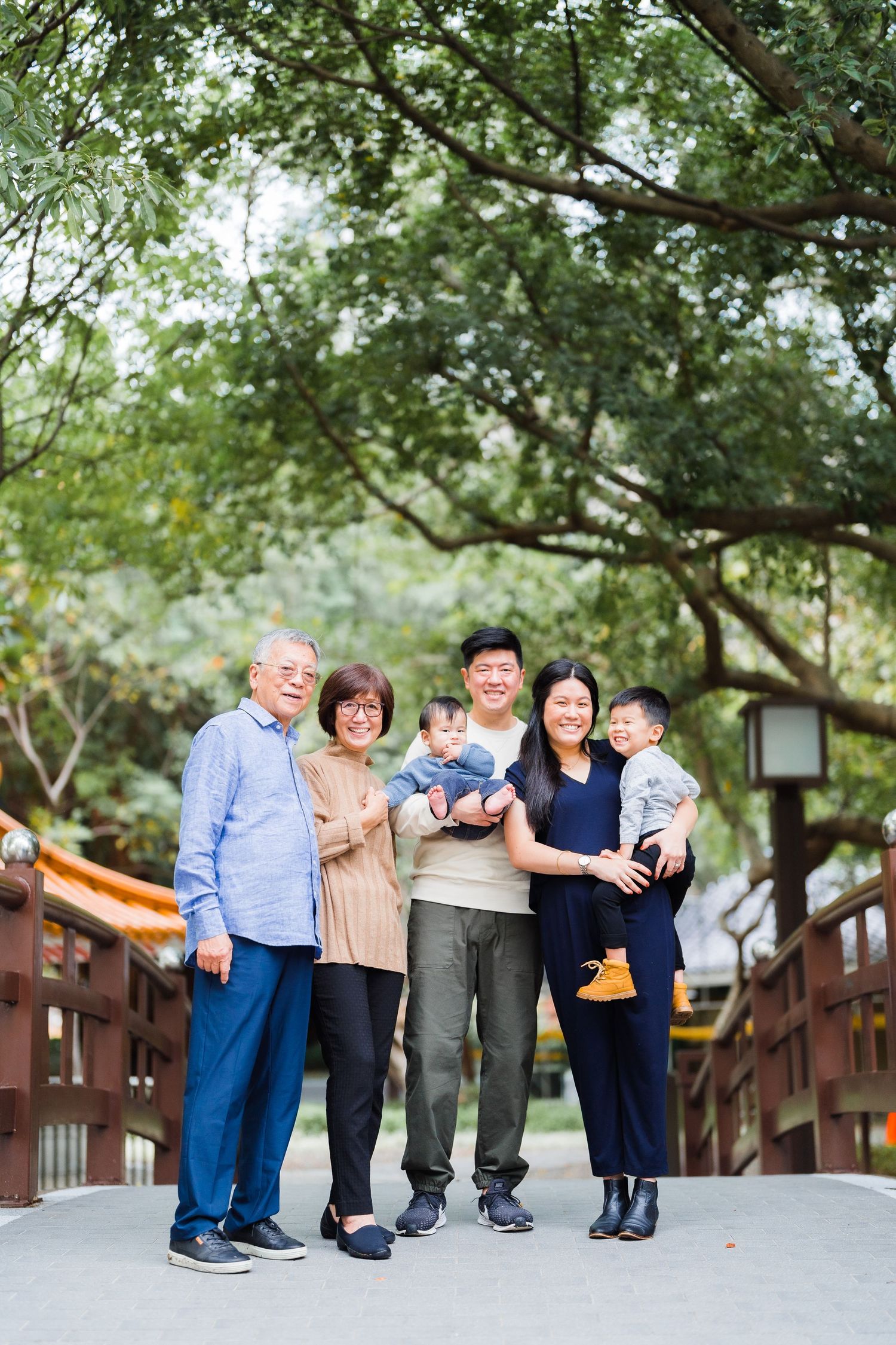 Multi-generational family standing together on wooden bridge in park setting.