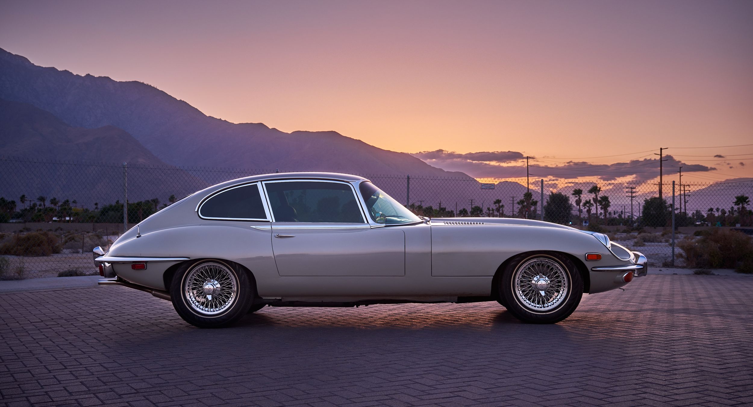 Silver Jaguar E-Type coupe silhouetted against purple sunset mountains.