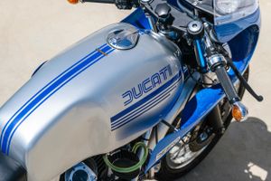 Close-up of a silver and blue Ducati motorcycle fuel tank with classic stripe design.