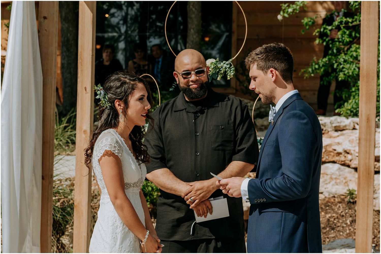 An intimate outdoor wedding ceremony moment between three people under a wooden arch.