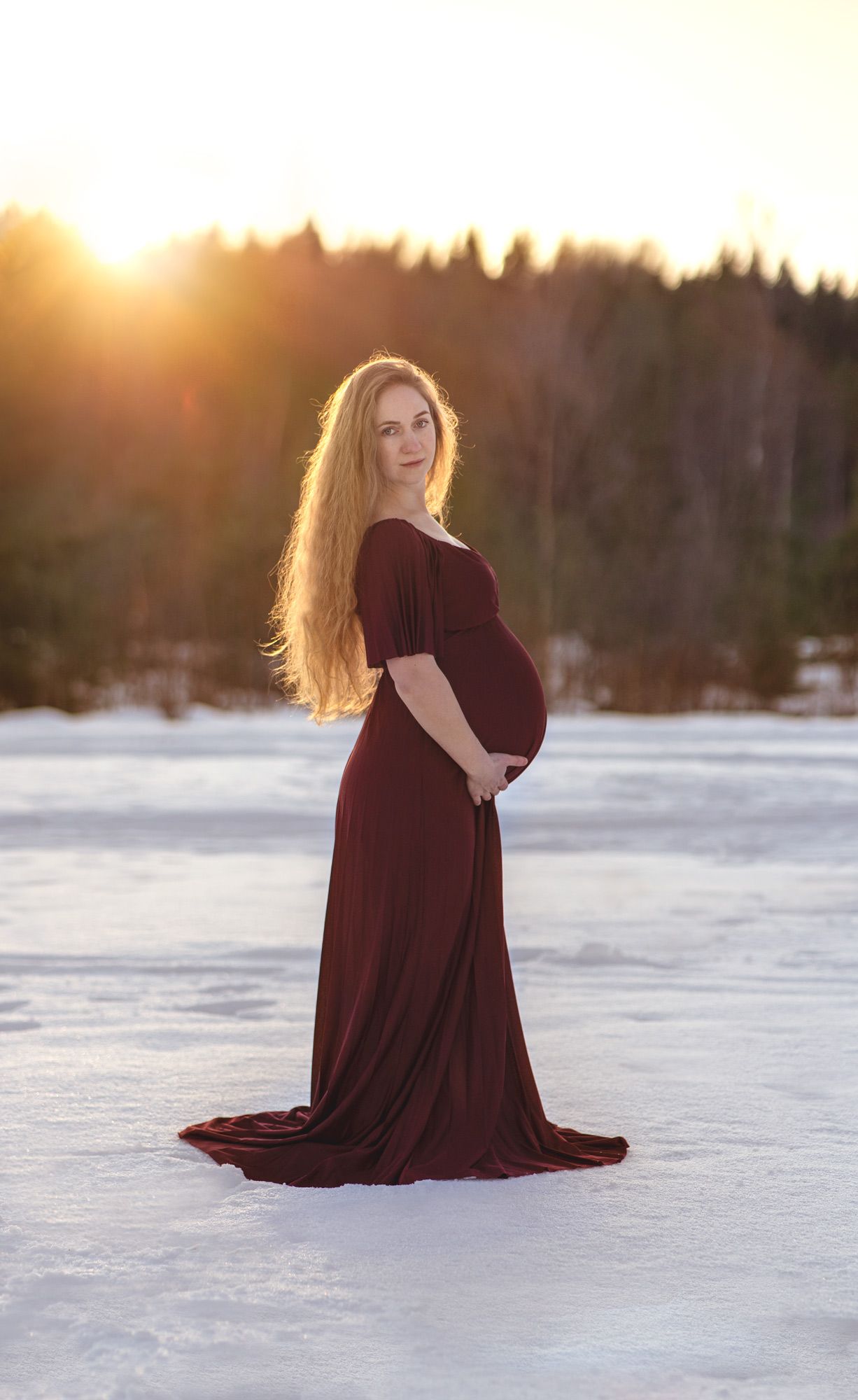 En gravid kvinna i vinröd långklänning håller om sin gravidmage i soluppgången. Det är en vintermorgon.