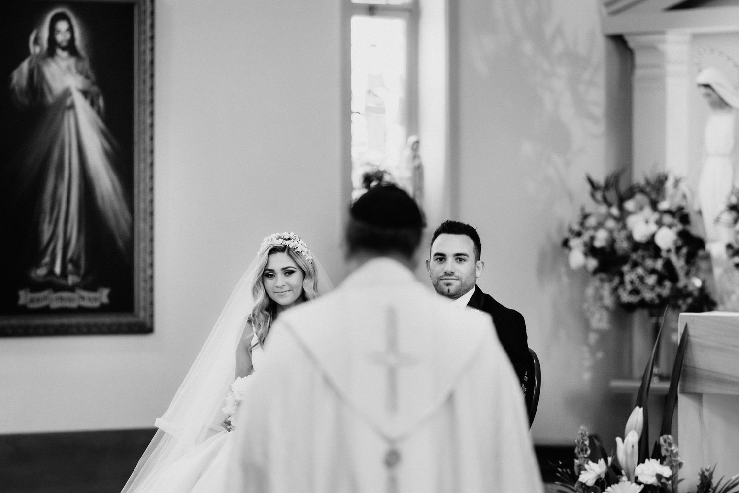 Black and white wedding ceremony photo of couple at church altar with priest and religious artwork.