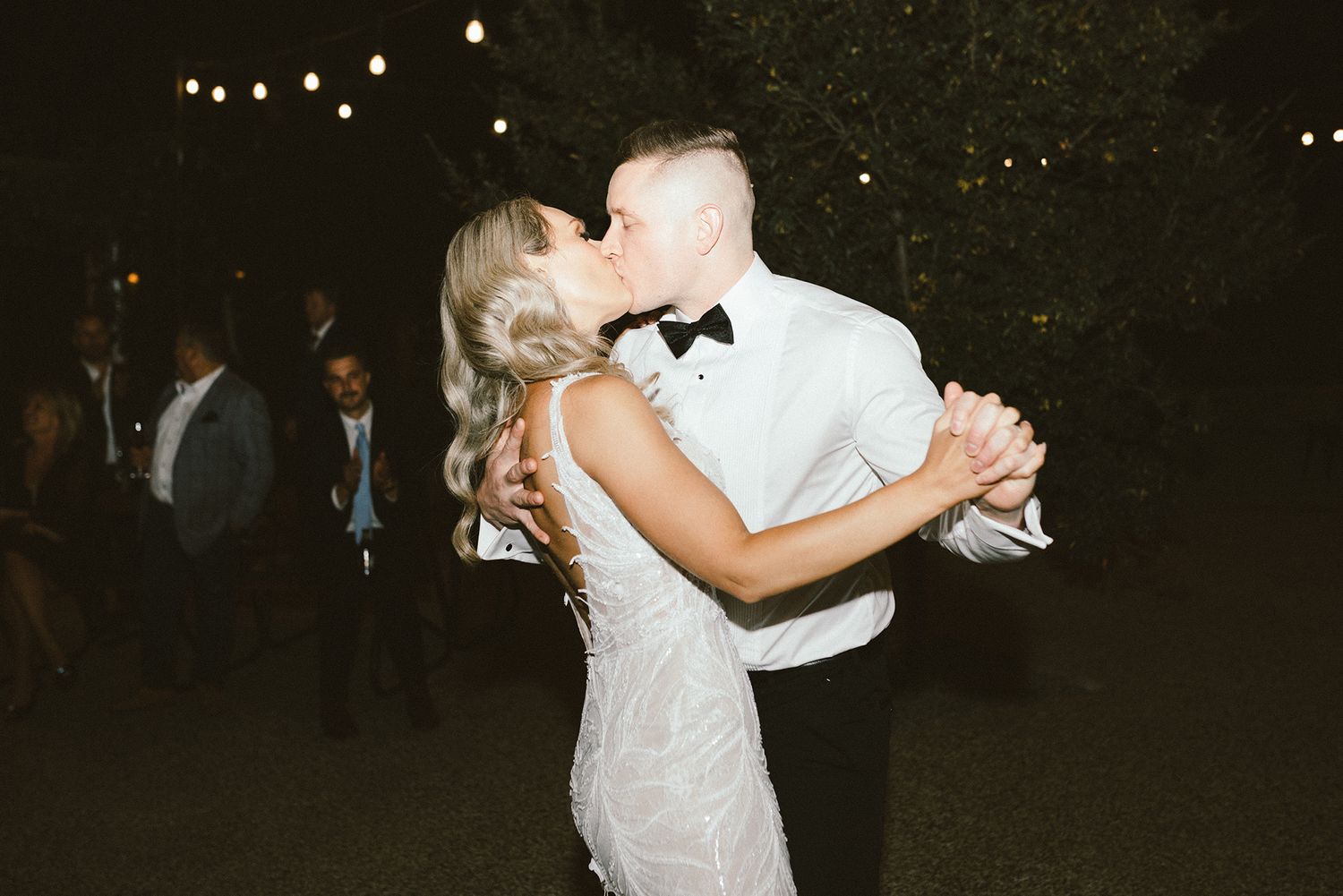 Wedding couple shares their first dance under twinkling outdoor lights at night.