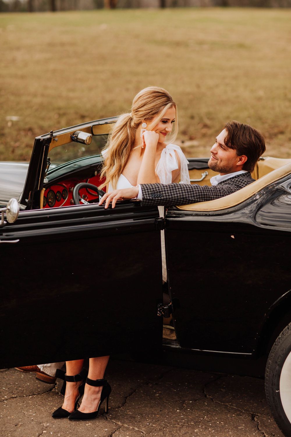 Bride in white dress and groom in plaid suit sharing romantic moment beside classic black convertible car.