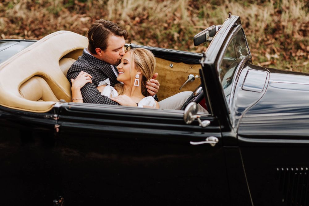 Couple sharing a tender kiss while seated in a vintage black convertible car surrounded by autumn scenery.