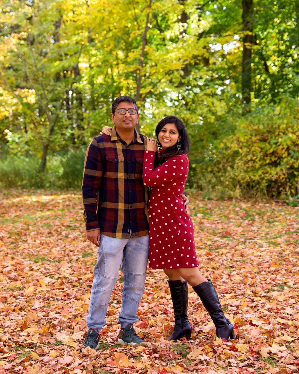 A couple poses together on fallen autumn leaves with colorful foliage in the background.