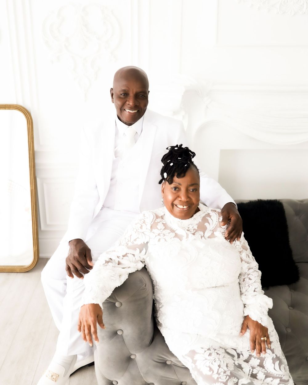 Couple in white formal attire posing on elegant grey chair.