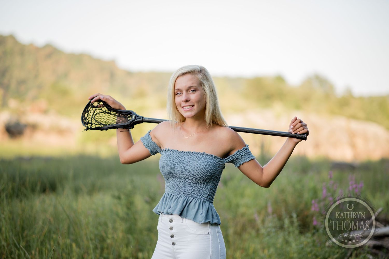 Woman in denim top poses with lacrosse stick against natural mountain backdrop.