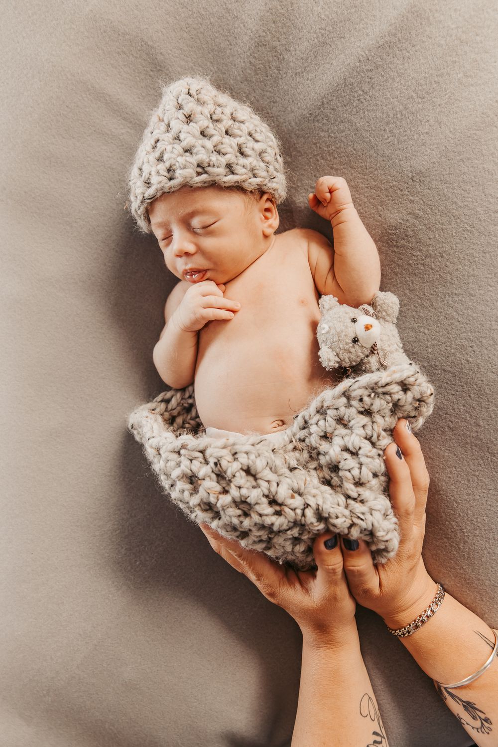 baby sleeping on grey blankets with a beanie and mums hands