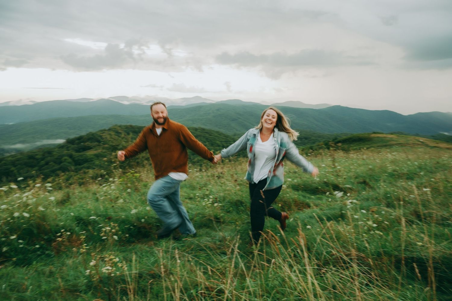 A couple runs joyfully through a grassy mountain meadow with scenic mountain views in the background.