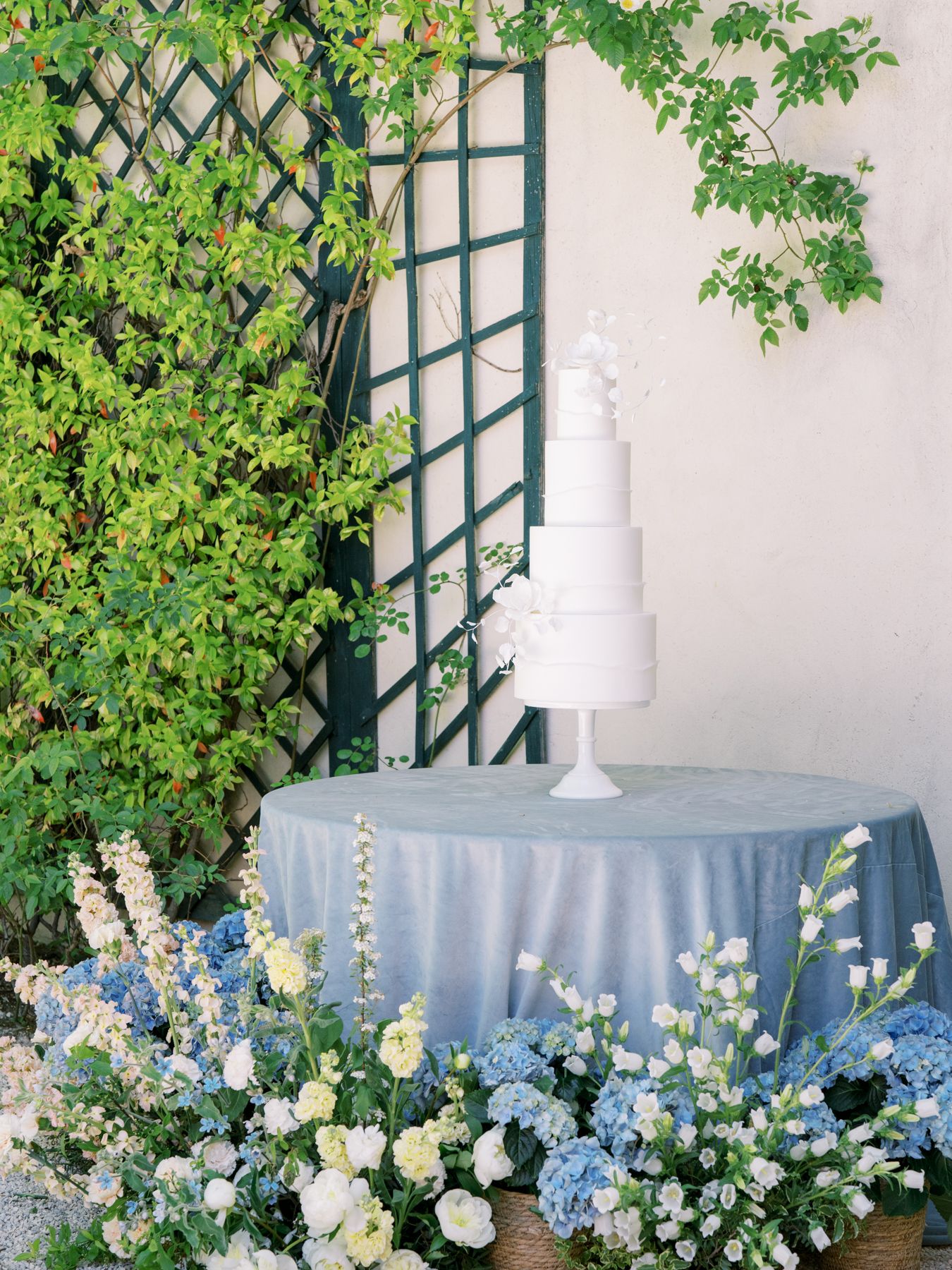 An elegant wedding cake display with blue tablecloth and floral arrangements against a garden trellis.