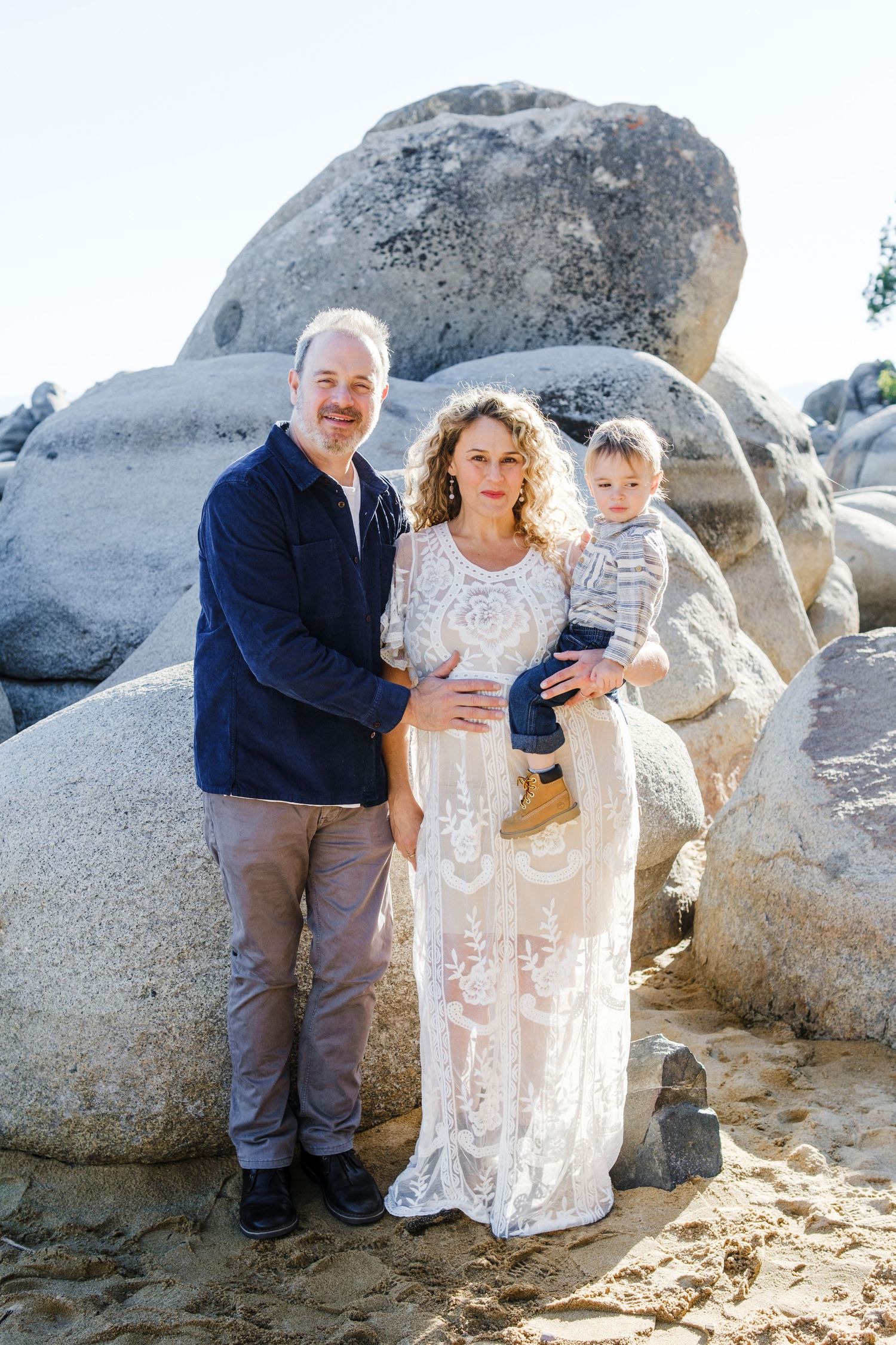 A family stands together posing by large coastal rocks on a sunny beach day.