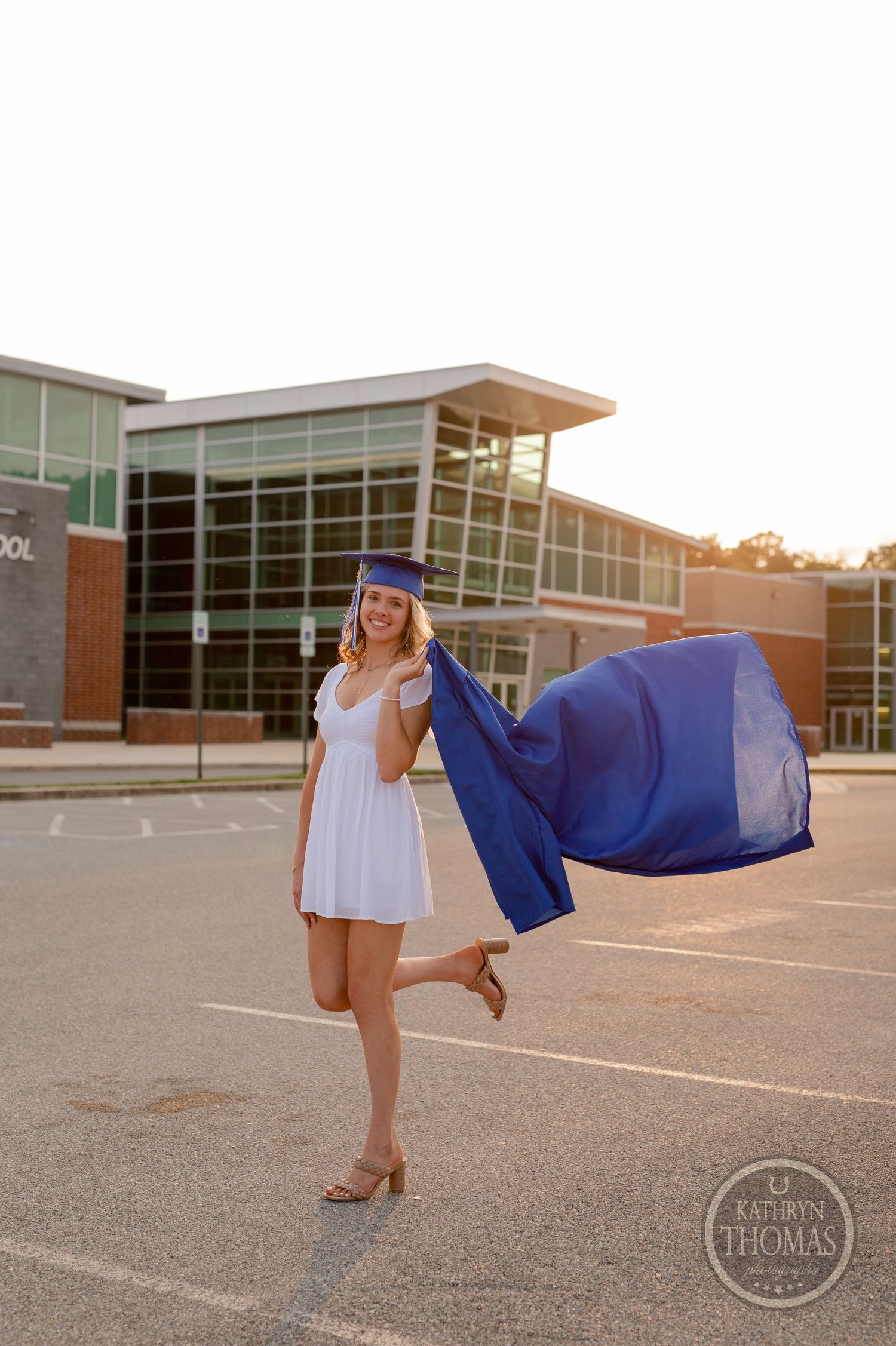 Graduate in blue cap and gown twirling in parking lot during golden hour.
