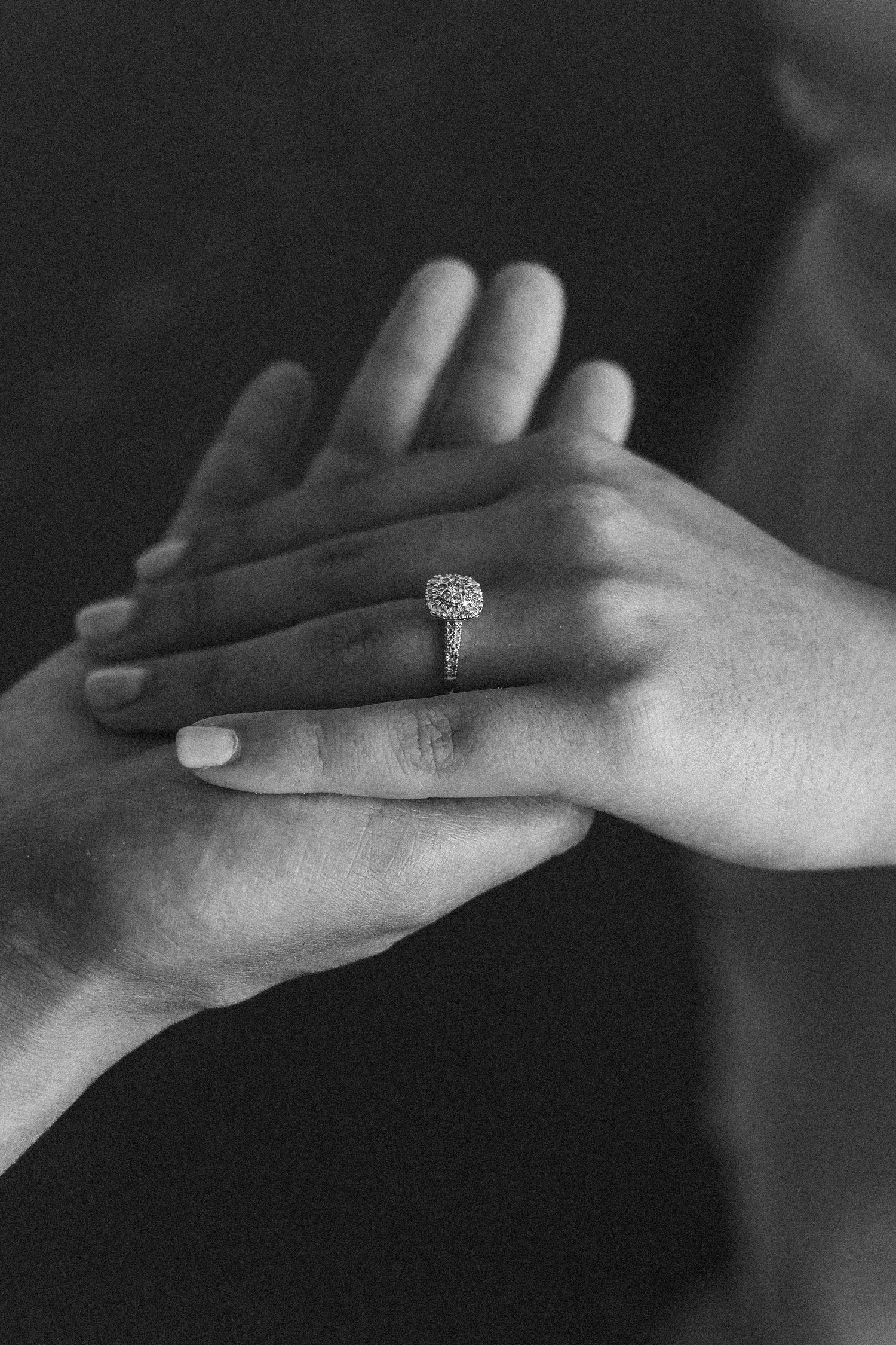 Black and white close-up of two hands touching, featuring a sparkling cushion-cut diamond engagement ring.